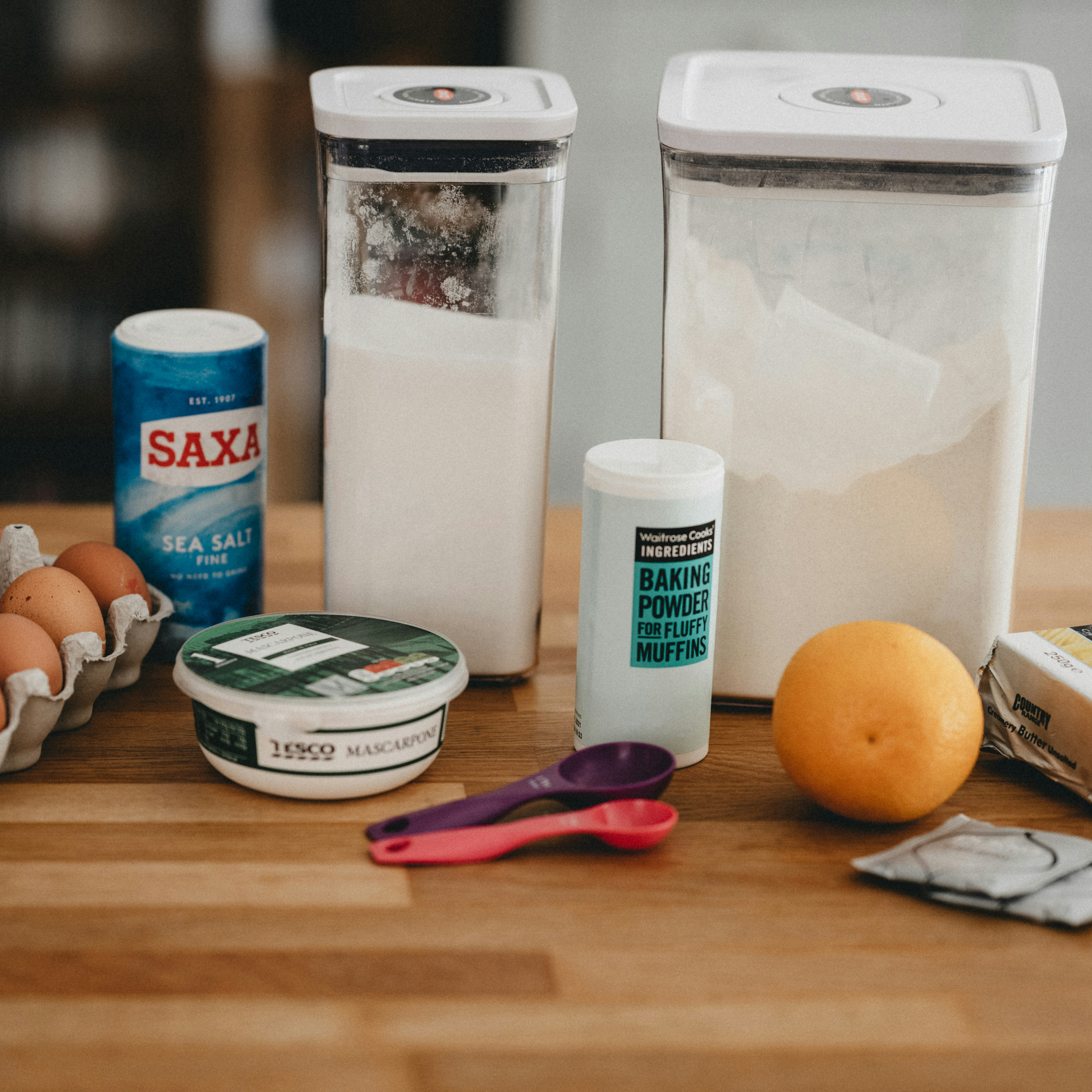 Eggs, salt, baking powder, butter, an orange, 2 containers of white powder, and 2 measuring spoons on a wooden countertop